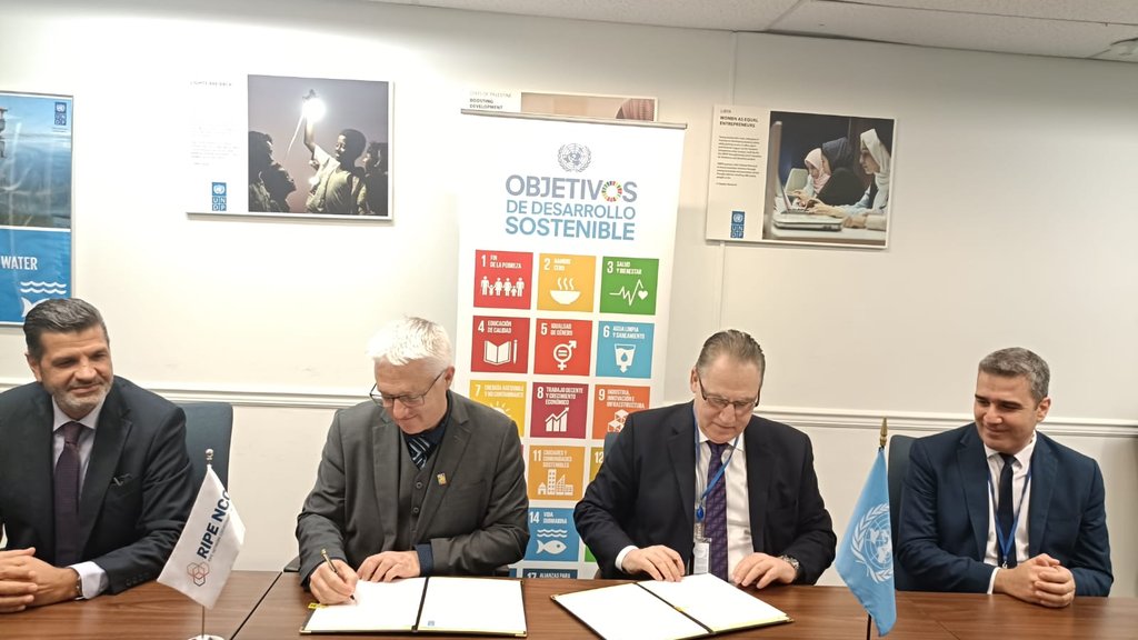 Four men sit at a brown table, two are signing an MoU document.